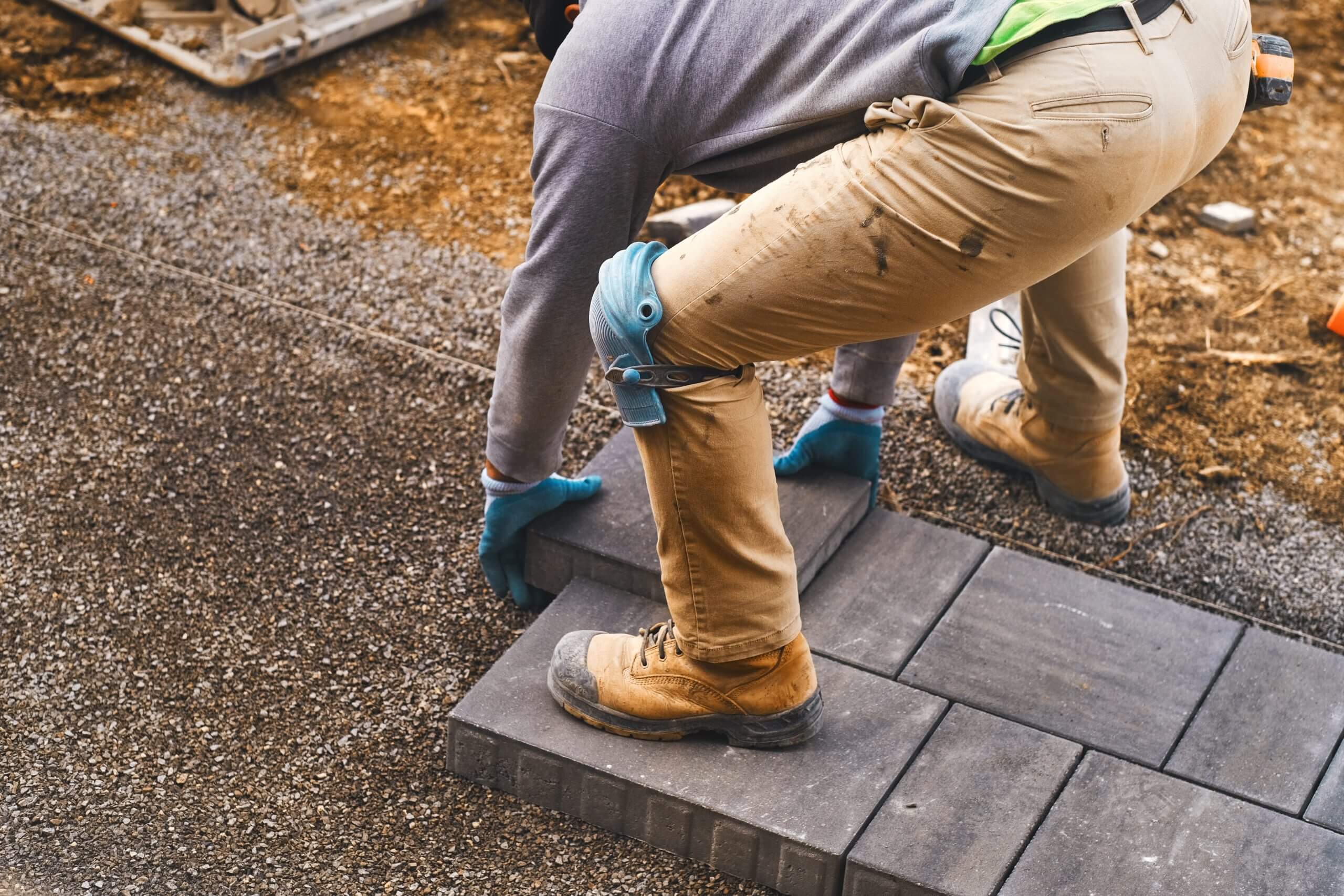 Landscaping paver worker laying paving stones on sandy ground of construction patio yard site in spring summer. Contractor wearing safety protective cloth, gloves and knee pads for installation work.